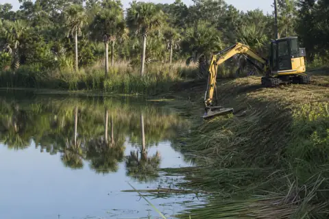 Excavator clearing woody growth from a pond bank while preserving the bank slope